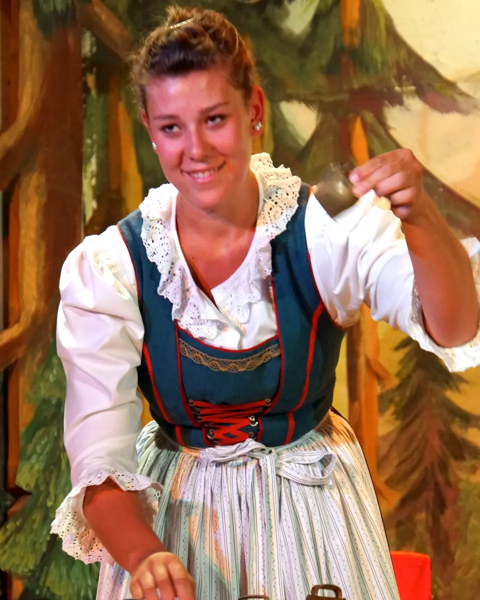 Žena v dirndlu rozeznívá řadu kravských zvonců položených na červenobíle kostkovaném stole před malovanými jedlemi