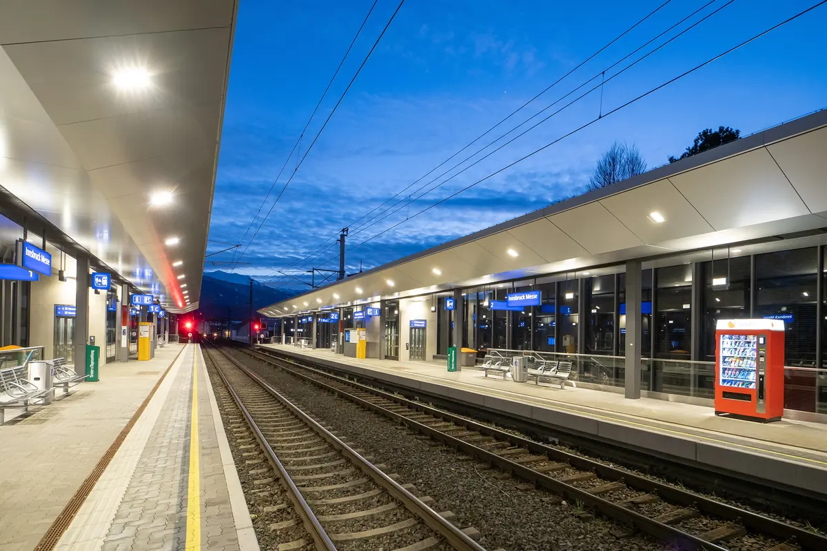 Nástupiště na zastávce Innsbruck Messe pod hluboce modrou večerní oblohou, podél kolejí osvětlené přístřešky