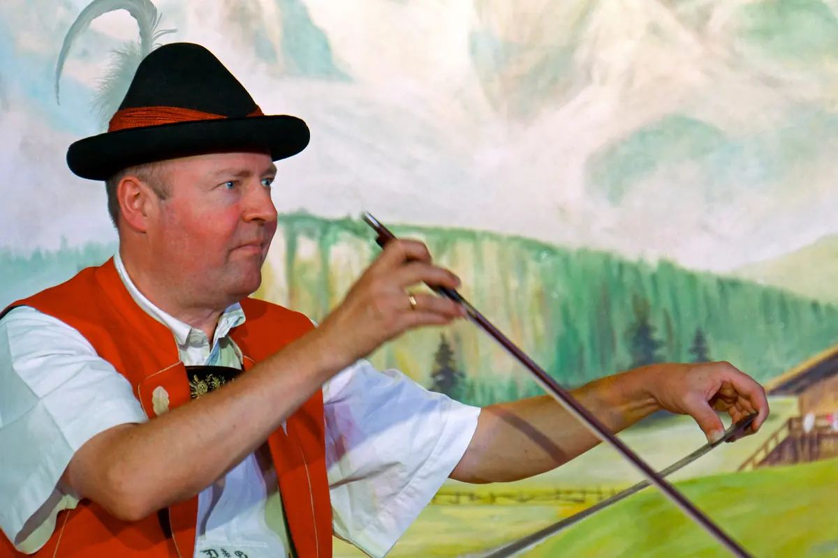 A man in a red waistcoat and a feathered hat draws a bow across a singing saw in front of a painted alpine meadow and cliffs