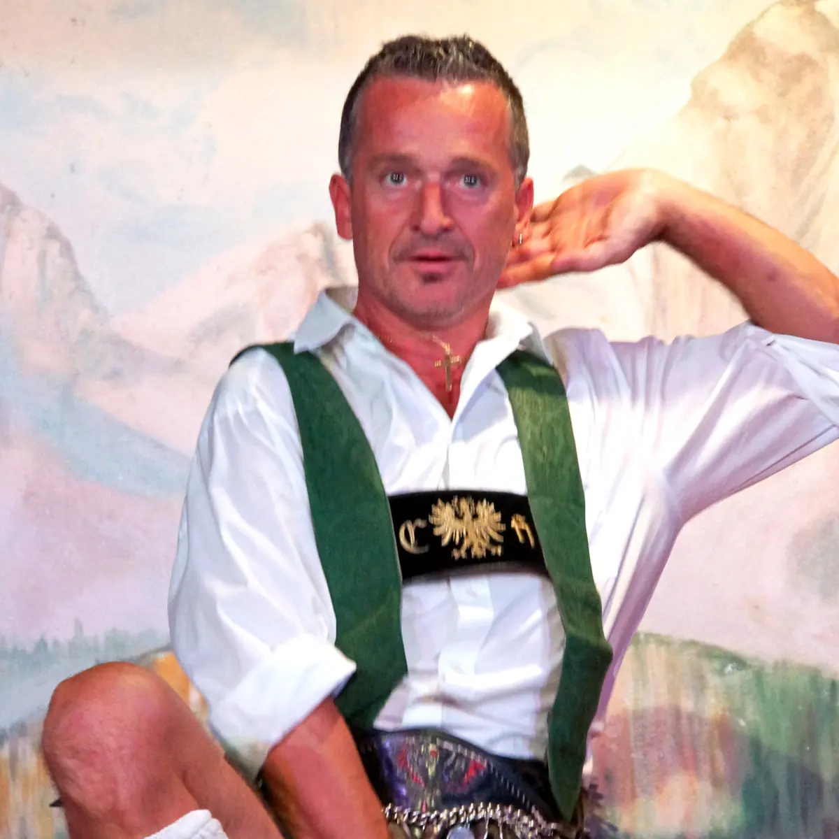 Muž v lederhosen a zelených šrácích tančí s jedním kolenem zvednutým a rukou za hlavou před malovanou horskou kulisou